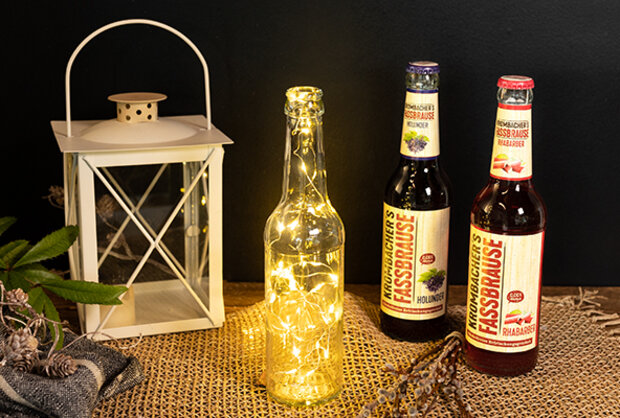 Eine leere Glasflasche mit Lichtern ist neben zwei Flaschen 'Fassbrause' von 'Fassbrause' platziert, eine in Holunder- und die andere in Rhabarbergeschmack. Im Hintergrund steht eine Laterne auf einem Jutesack.
