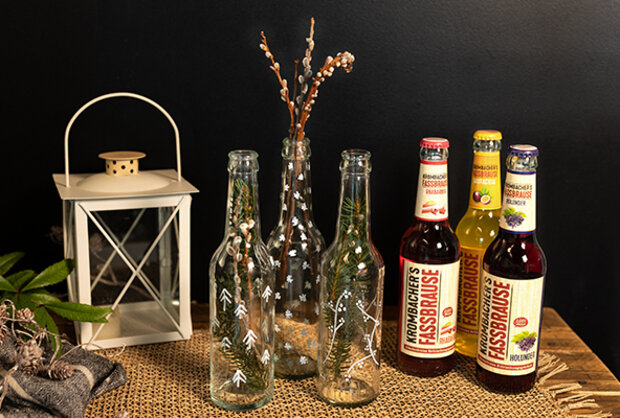 Drei Flaschen mit verschiedenen Designs und zwei Flaschen 'Krahnbrichers Fassbrause' in rot und gelb stehen auf einem Tisch. Im Hintergrund befindet sich eine Laterne und eine Vase mit Zweigen.