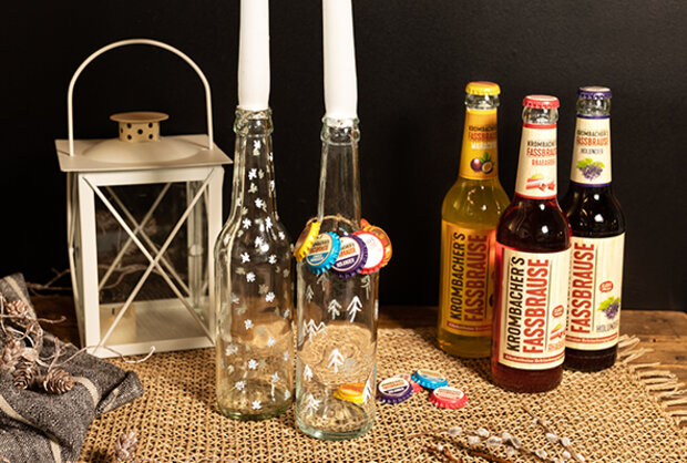 Zwei leere Flaschen mit bunten Verschlüssen und einem dekorativen Muster stehen zwischen drei Flaschen Kronenbacher's Fassbrause in verschiedenen Farben. Im Hintergrund befindet sich eine weiße Laterne mit einem Holzdeckel.