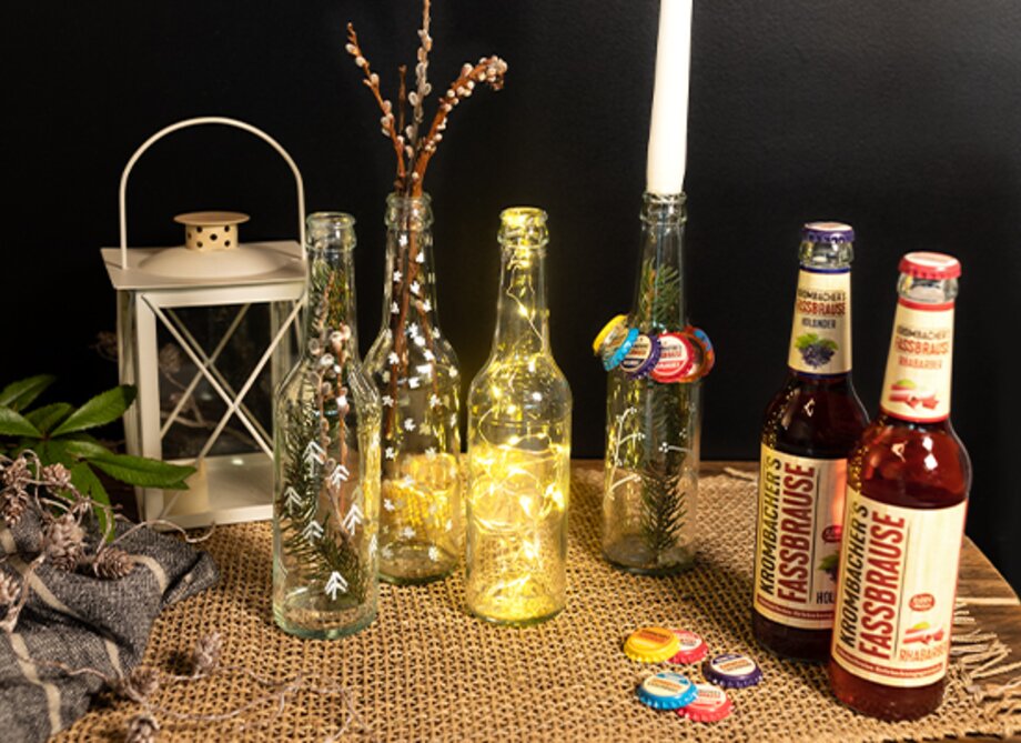 Zwei Flaschen KISSBRÄUSE stehen neben einer leuchtenden Flasche und einer Laterne auf einem Tisch. Im Hintergrund sind dekorative Zweige und Kronkorken sichtbar.