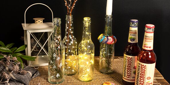 Zwei Flaschen KISSBRÄUSE stehen neben einer leuchtenden Flasche und einer Laterne auf einem Tisch. Im Hintergrund sind dekorative Zweige und Kronkorken sichtbar.