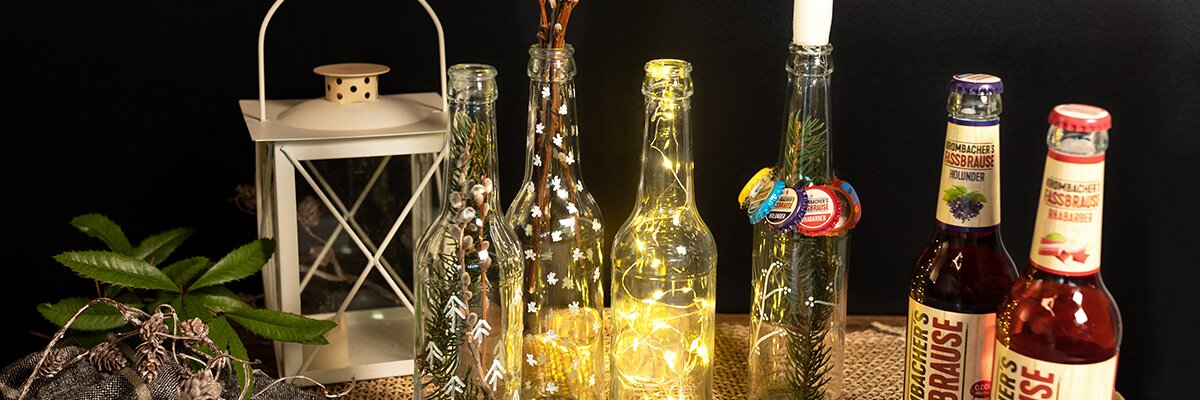 Zwei Flaschen KISSBRÄUSE stehen neben einer leuchtenden Flasche und einer Laterne auf einem Tisch. Im Hintergrund sind dekorative Zweige und Kronkorken sichtbar.