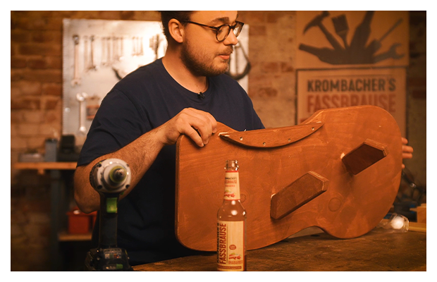 Mann stellt ein Holzbrett auf den Tisch und hält es fest. Auf dem Tisch ist auch eine Fassbrause.