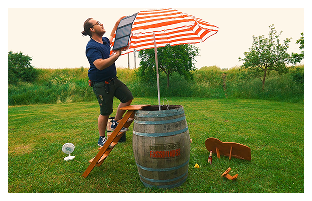 Ein Mann auf einer Leiter bringt Solarpanel auf einem Sonnenschirm an, der auf einem Holzfass gesteckt ist.