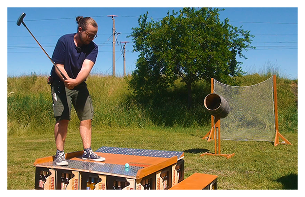Ein aufgetselltes Fass auf einer Holzstange wurde aufgstellt und dient als Ziel für einen Golfschlag. Ein Mann steht auf einer Erhöhung mit einem Golfschläger