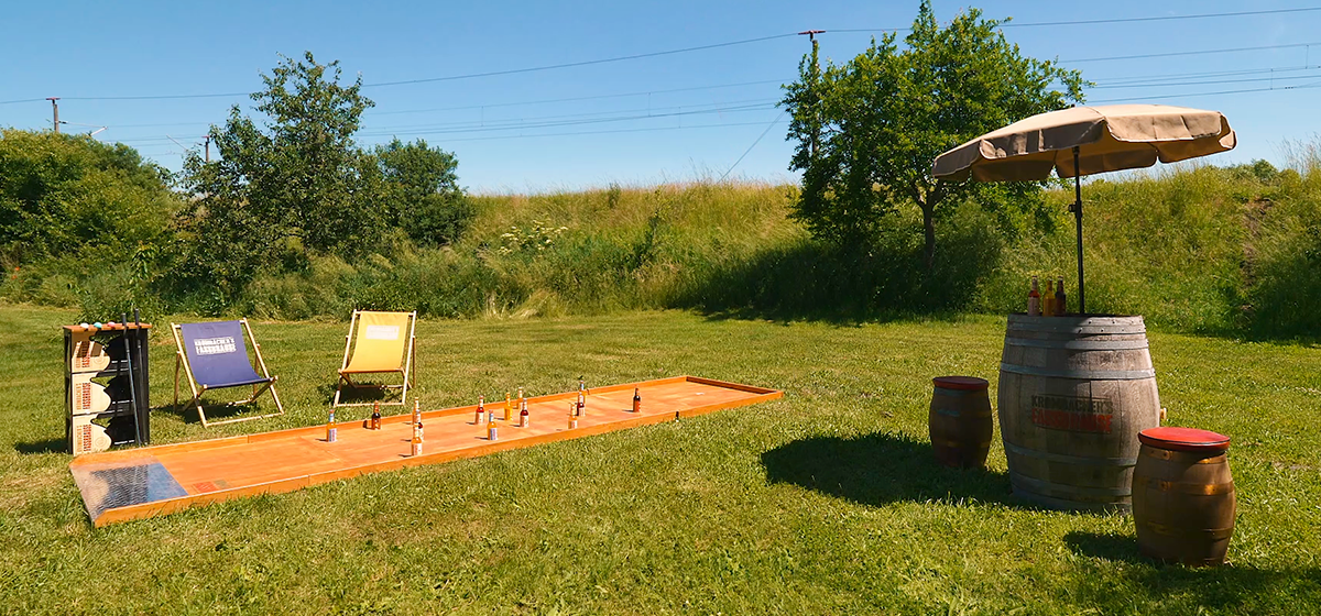 Zwei Sommerklappstühle und eine Holzgolfbahn mit Krombacher Fassbrauseflaschen als Hindernis sind auf einer Wiese aufgestellt