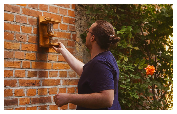 Das Vogelhäuschen aus Holz mit einer gefüllten Glasflasche mit Vogelfutter wird an eine Backsteinwand gehangen.