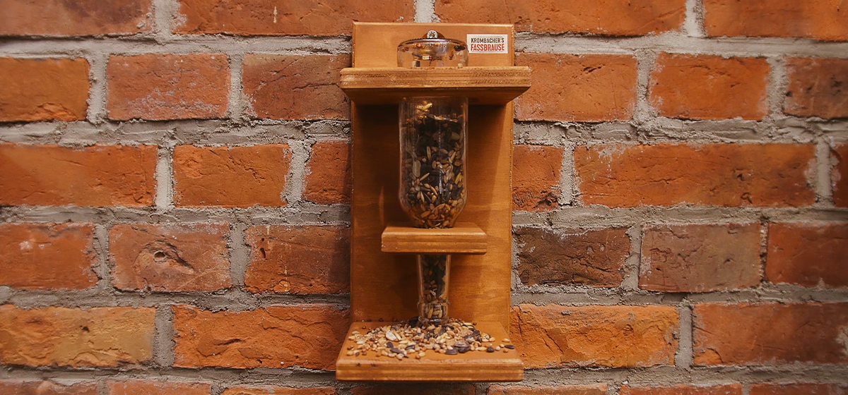 Eine Glasflasche steckt in einem Holzständer und ist mit Vogelfutter gefüllt.