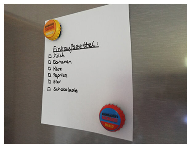 weißer Zettel mit Text: Einkaufszettel und Unterpunkten, die mit zwei Kronkorkenmagneten gehalten werden