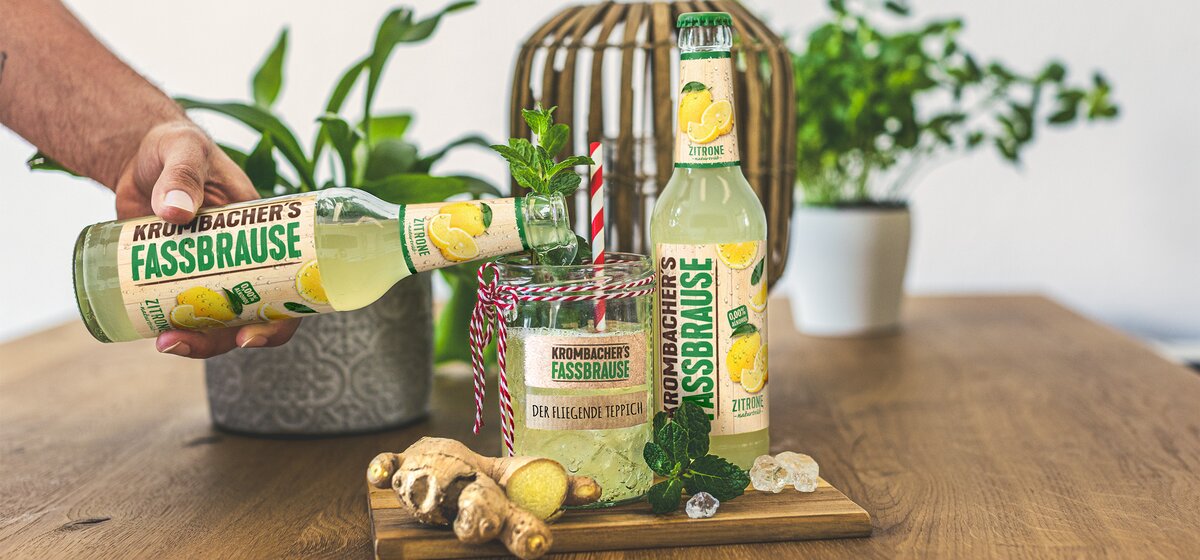 Eine Hand hält eine Flasche 'Krumbacher's Fassbrause' mit Zitrone, während eine zweite Flasche und ein Glas mit einem Getränk auf einem Holztisch stehen. Das Glas ist mit einem Strohhalm und frischen Minzblättern dekoriert.