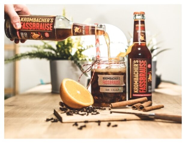 Eine Person gießt Krombacher's Fassbrause aus einer Flasche in ein Glas mit einer Orangenscheibe und Zimtstangen. Im Hintergrund sind Pflanzen und eine Lampe sichtbar.