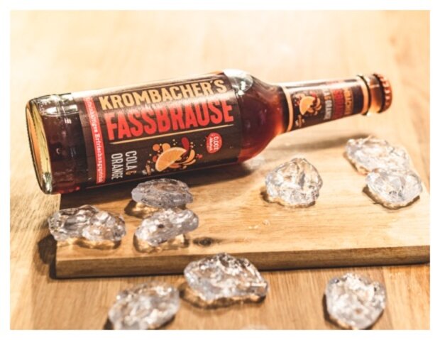 Eine Flasche Krombacher's Fassbrause Cola-Orange liegt auf einem Holzbrett, umgeben von Eiswürfeln. Die Flasche ist rot und zeigt das Logo sowie den Produktnamen.