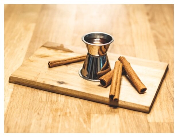 Ein Edelstahlmessbecher steht auf einem Holzbrett, umgeben von mehreren Zimtstangen. Die Zimtstangen sind in verschiedenen Längen angeordnet.