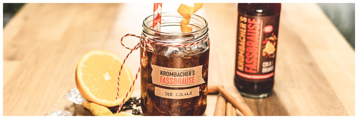 Ein Glas mit Krombacher's Fassbrause, dekoriert mit einer Orangenscheibe und zwei Strohhalmen, steht auf einem Holztisch. Im Hintergrund ist eine Flasche Krombacher's Fassbrause sichtbar.