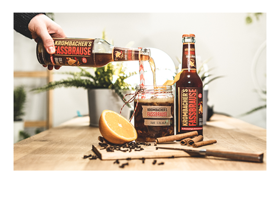 Eine Hand gießt Krombacher's Fassbrause in ein Glas mit Zimt und einer Orangenscheibe. Auf dem Tisch stehen eine Flasche Fassbrause und eine weitere Flasche mit ähnlichem Design.