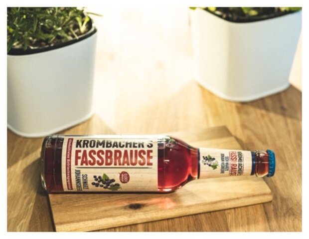 Eine Flasche Krombacher's Fassbrause liegt auf einem Holzbrett. Im Hintergrund sind zwei weiße Blumentöpfe mit Pflanzen sichtbar.