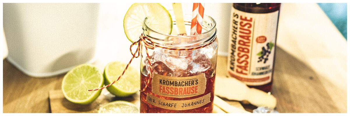 Ein Glas mit Krombacher's Fassbrause, gefüllt mit einem roten Getränk, dekoriert mit einer Limettenscheibe und einem Strohhalm. Im Hintergrund steht eine Flasche Krombacher's Fassbrause und frische Limetten.