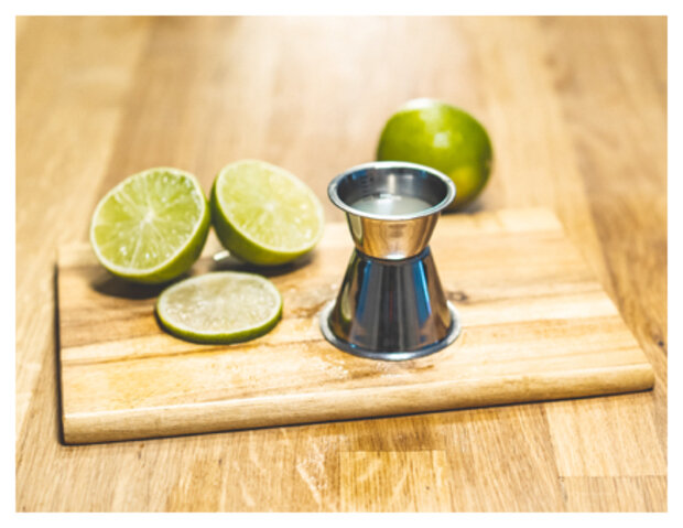 Ein Messbecher aus Edelstahl steht auf einem Holzbrett, umgeben von zwei halbierten Limetten und einer ganzen Limette. Die Limetten sind grün und frisch.