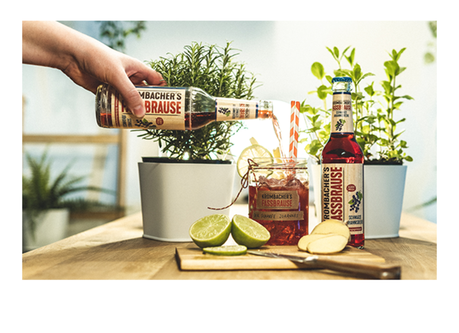 Eine Hand gießt 'Krombacher's Fassbrause' in ein Glas mit Limetten und Ingwer. Im Hintergrund stehen zwei Pflanzen in Töpfen.