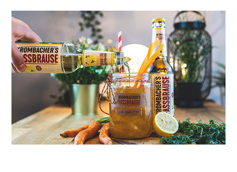 Eine Person gießt Krombacher's Fassbrause in ein Glas mit einer orangefarbenen Flüssigkeit, garniert mit Karotten und Zitronenscheiben. Im Hintergrund stehen zwei Flaschen Krombacher's Fassbrause und Pflanzen.