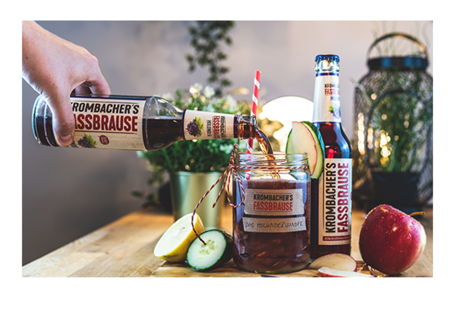 Eine Hand gießt Krombacher's Fassbrause aus einer Flasche in ein Glas. Auf dem Tisch liegen ein Apfel und geschnittene Äpfel neben der Getränkeflasche.