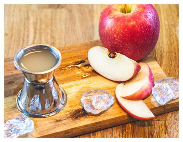 Ein silberner Messbecher steht auf einem Holzbrett neben einem roten Apfel und geschnittenen Apfelstücken. Um den Messbecher sind einige Eiswürfel angeordnet.