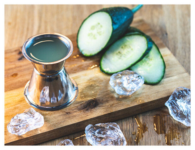 Ein silberner Becher mit grünem Getränk steht auf einem Holzbrett neben geschnittenen Gurkenscheiben. Um den Becher liegen einige Eiswürfel und ein wenig Honig.