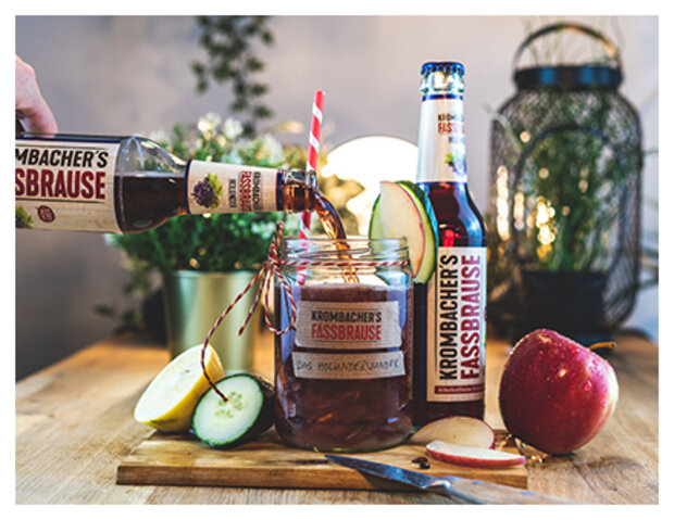 Eine Flasche Krombacher's Fassbrause wird in ein Glas mit einer roten und weißen gestreiften Strohhalm gegossen. Neben dem Glas stehen eine Flasche Fassbrause, ein roter Apfel und Apfelscheiben.
