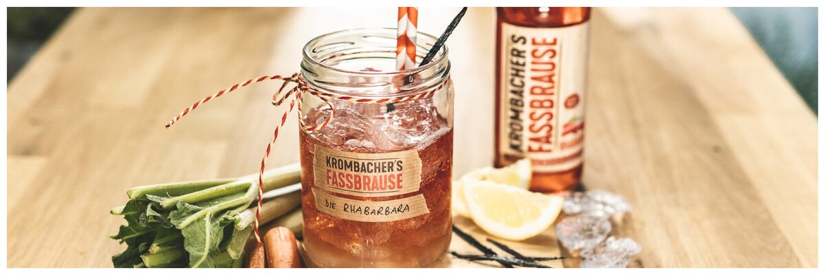 Ein Glas mit Krombachers Fassbrause, gefüllt mit einem roten Getränk, Eiswürfeln und einem Strohhalm. Neben dem Glas stehen Sellerie, eine Zitronenscheibe und eine Flasche Krombachers Fassbrause.