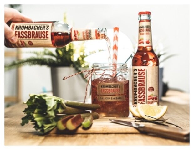 Eine Person gießt Krombacher's Fassbrause aus einer Flasche in ein Glas mit einem roten Strohhalm. Auf dem Tisch liegen Sellerie und eine Limette neben dem Glas.