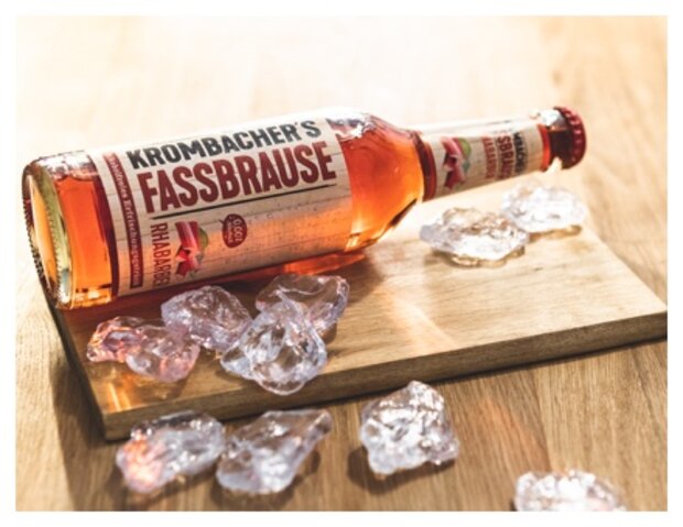 Eine Flasche Krombacher's Fassbrause mit Rhabarbergeschmack liegt auf einem Holzbrett, umgeben von zerstoßenem Eis. Die Flasche ist in einer schrägen Position und zeigt das Etikett deutlich.
