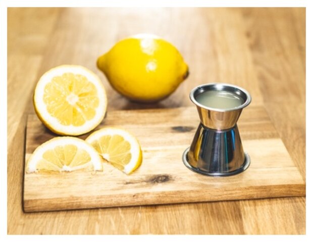 Eine ganze Zitrone und zwei halbierte Zitronen liegen auf einem Holzbrett. Neben den Zitronen steht ein Messbecher aus Edelstahl mit Zitronensaft.