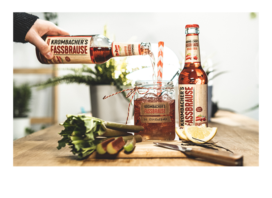 Eine Person gießt Krombacher's Fassbrause aus einer Flasche in ein Glas mit einem Strohhalm. Neben dem Glas stehen eine weitere Flasche Fassbrause, Zitronenscheiben und Selleriestangen.