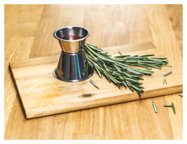Ein Edelstahl-Messbecher steht auf einem Holzbrett, umgeben von frischen Rosmarinzweigen. Der Messbecher enthält eine rote Flüssigkeit.