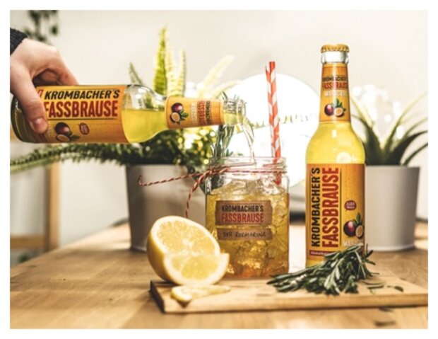 Eine Hand gießt Krombacher's Fassbrause aus einer Flasche in ein Glas mit einem Strohhalm. Auf dem Tisch liegen eine halbe Zitrone und frische Rosmarinzweige.