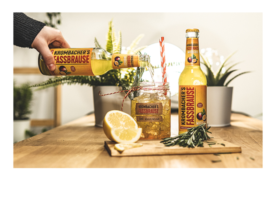 Eine Flasche Krombacher's Fassbrause wird über ein Glas mit einer Zitronenscheibe und einem Strohhalm gegossen. Im Hintergrund sind Pflanzen und eine Holzplatte sichtbar.