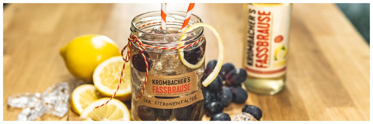 Ein Glas mit Krombacher's Fassbrause, gefüllt mit Eis, Trauben und Zitronenscheiben, steht auf einem Holztisch. Daneben befindet sich eine Flasche Krombacher's Fassbrause und eine Zitrone.