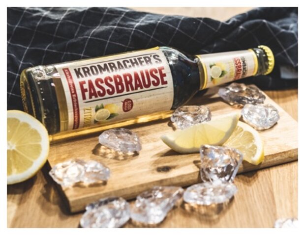 Eine Flasche Krombacher's Fassbrause liegt auf einem Holzbrett, umgeben von Eiswürfeln und Zitronenscheiben. Die Flasche hat ein etikettiertes Design mit dem Schriftzug 'Krombacher's Fassbrause'.