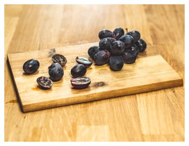 Ein Holzbrett mit einer Ansammlung von schwarzen Trauben und einigen aufgeschnittenen Trauben, die ihre innere Struktur zeigen. Die Trauben sind auf einer Holzoberfläche angeordnet.