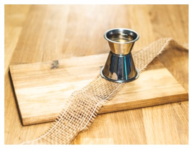 Ein glänzender, silberner Messbecher steht auf einem Holzbrett. Daneben liegt ein Stück aus Jute gewebtes Material.