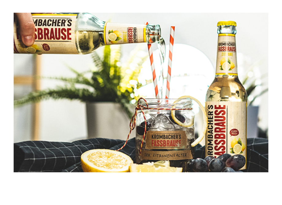 Eine Person gießt Krombacher's Fassbrause in ein Glas mit Eis und Zitronenscheiben. Neben dem Glas stehen zwei Flaschen Krombacher's Fassbrause.