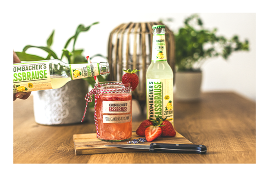 Eine Hand hält eine Flasche Krombacher's Fassbrause über einem Glas mit rotem Getränk, das mit einem Etikett 'Der Gartenzauberer' versehen ist. Im Hintergrund stehen zwei Flaschen Fassbrause und eine Erdbeeren auf einem Holzbrett.