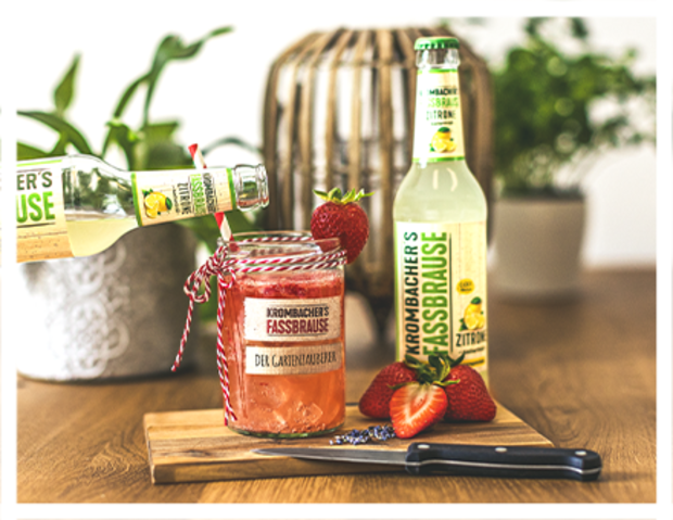 Eine Hand hält eine Flasche Krombacher's Fassbrause über einem Glas mit rotem Getränk, das mit einem Etikett 'Der Gartenzauberer' versehen ist. Im Hintergrund stehen zwei Flaschen Fassbrause und eine Erdbeeren auf einem Holzbrett.