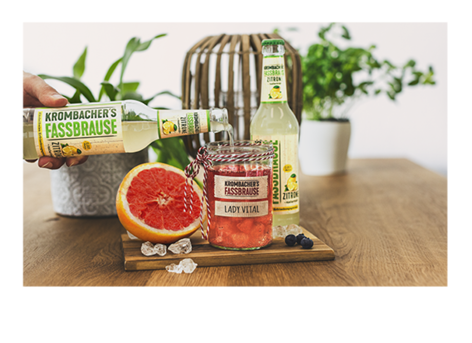 Eine Hand hält eine Flasche Krombacher's Fassbrause über einem Glas mit rotem Getränk, das mit einem Etikett 'LADY VITAL' versehen ist. Im Hintergrund stehen zwei Flaschen Fassbrause und eine Grapefruit auf einem Holzbrett.
