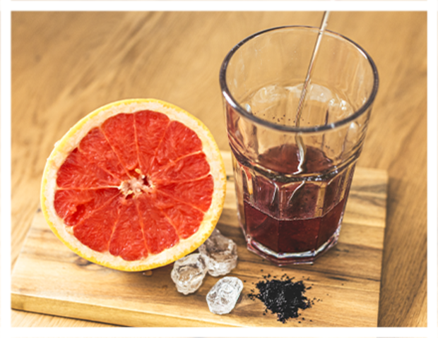 Eine halbierte Grapefruit liegt auf einem Holzbrett neben einem Glas mit rotem Saft und Eiswürfeln. Ein Rührstab ist im Glas sichtbar.