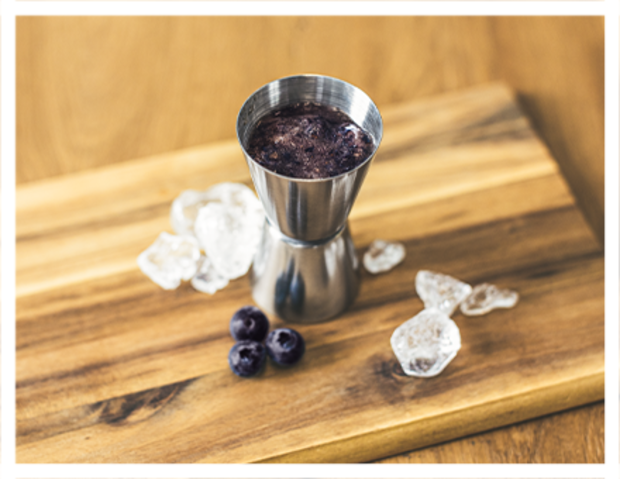 Ein Edelstahl-Jigger steht auf einem Holzbrett, umgeben von Eiswürfeln und drei Blaubeeren. Der Jigger enthält eine dunkle Flüssigkeit.
