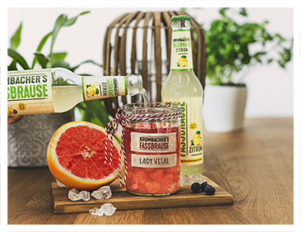 Zwei Flaschen 'Krombacher's Fassbrause' stehen neben einem Glas mit rotem Getränk, das mit einer roten Schnur dekoriert ist. Auf dem Glas steht 'LADY VITAL', daneben liegen Eiswürfel und eine Grapefruit.