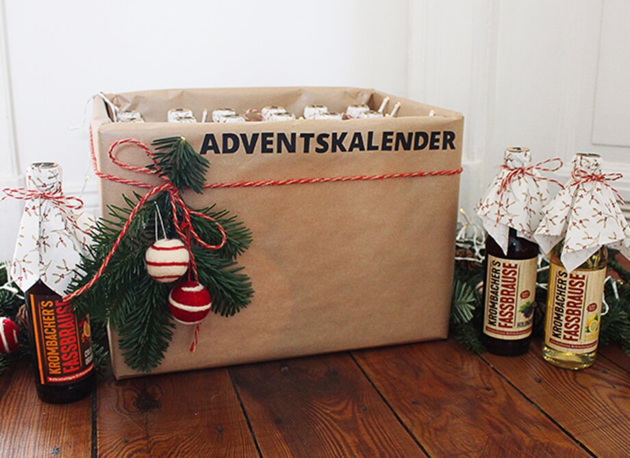 Ein Adventskalender aus braunem Papier mit der Aufschrift 'ADVENTSKALENDER' und dekoriert mit Tannenzweigen und roten Kugeln. Neben dem Kalender stehen zwei Flaschen mit dem Etikett 'Kronbacher's Kessbräuse'.