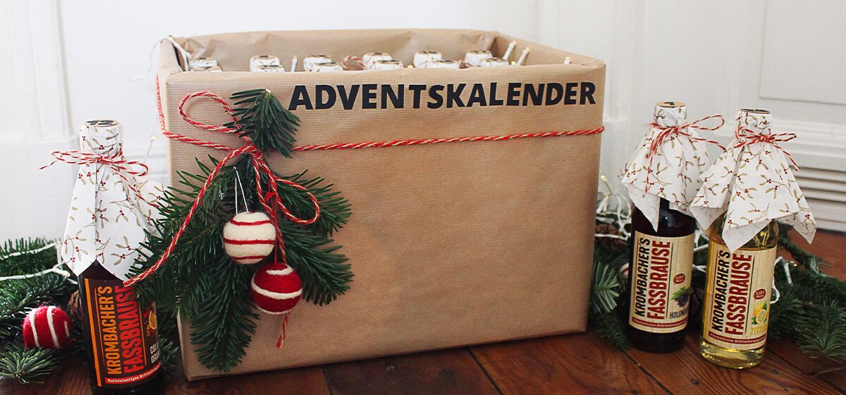 Ein Adventskalender aus braunem Papier mit der Aufschrift 'ADVENTSKALENDER' und dekoriert mit Tannenzweigen und roten Kugeln. Neben dem Kalender stehen zwei Flaschen mit dem Etikett 'Kronbacher's Kessbräuse'.