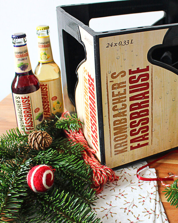 Ein Kasten mit 'IRONBACHER'S FASSBRAUSE' steht auf einem Tisch, umgeben von zwei Flaschen in rot und gelb. Dekorative Tannenzweige und eine rote Kugel sind ebenfalls sichtbar.